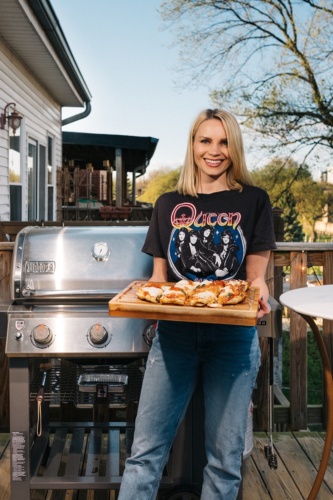 blonde woman holding a grilled pizza in front of a grill